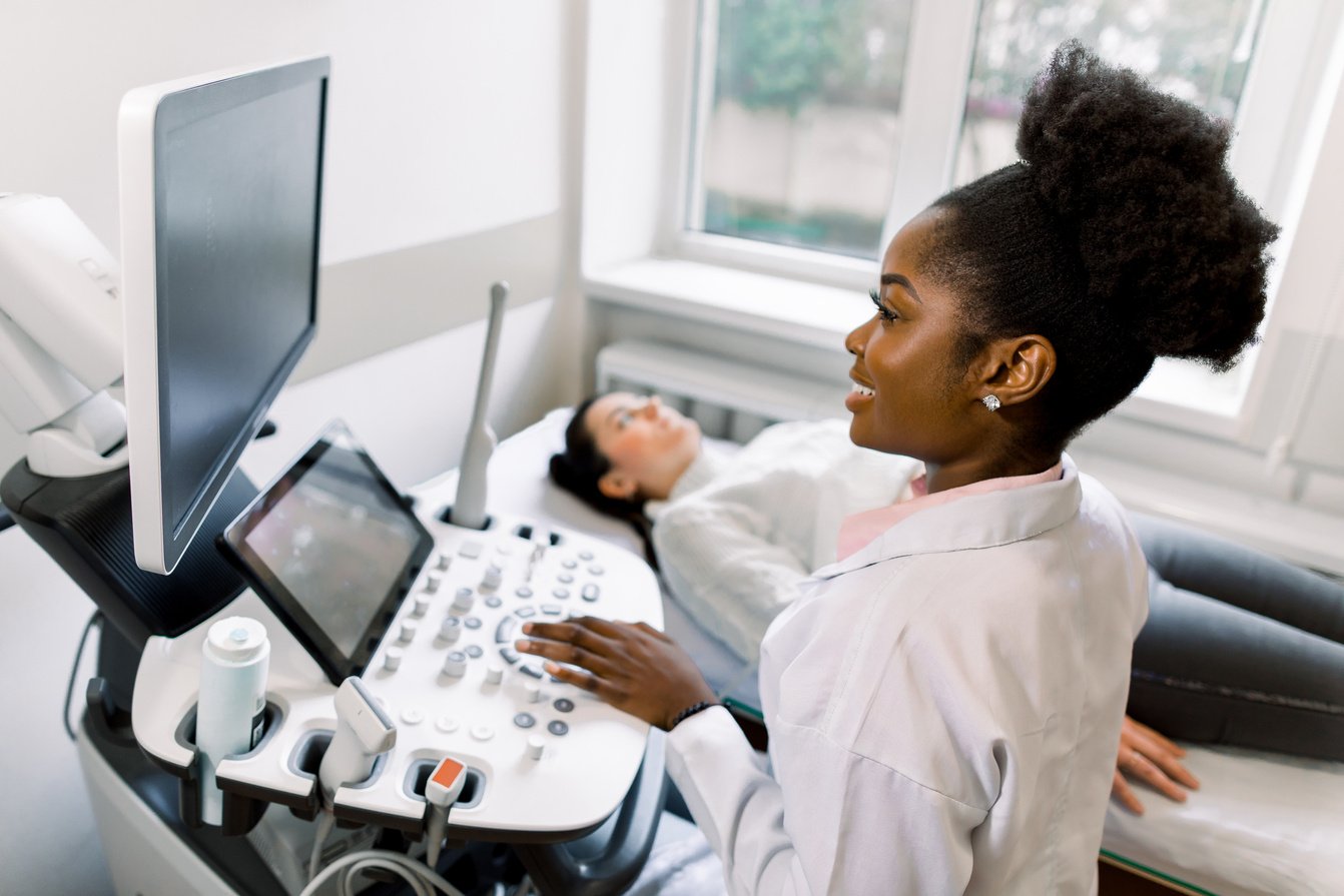 Young African female doctor using ultrasound machine in clinic, makes abdominal ultrasound for pregnant woman patient. Ultrasound Scanner. Sonography.